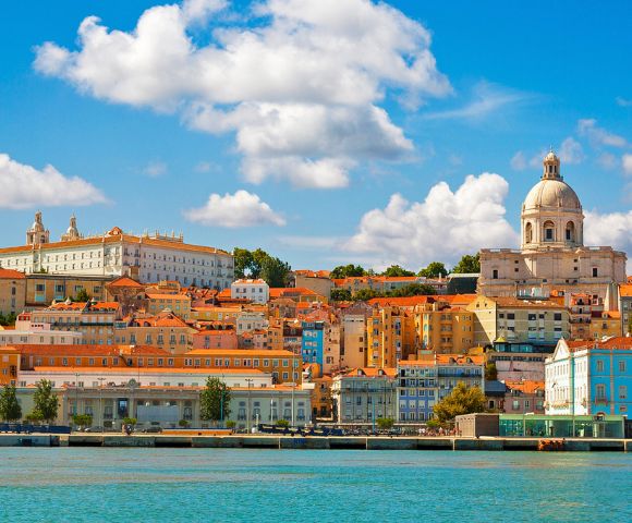 Scenic view of Lisbon with colorful buildings and a domed cathedral under a blue sky with fluffy clouds, reflecting on the calm waterfront.