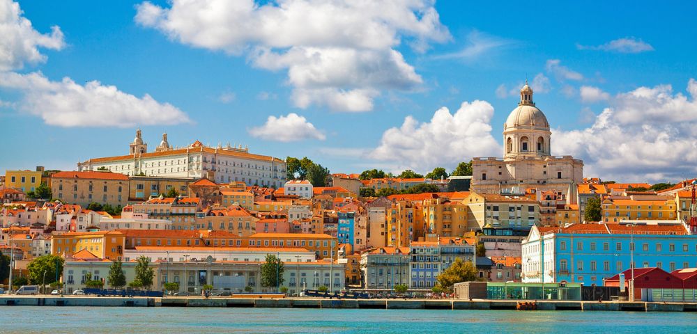 Scenic view of Lisbon with colorful buildings and a domed cathedral under a blue sky with fluffy clouds, reflecting on the calm waterfront.