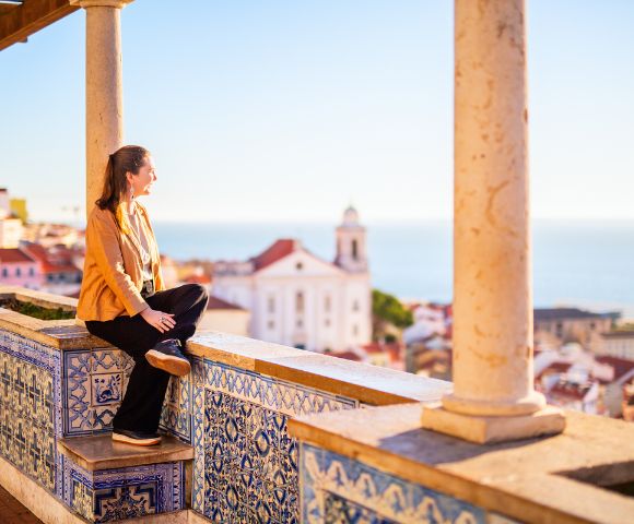 Woman in a yellow jacket sits on a tiled terrace, gazing at a scenic coastal town with a white church and red rooftops under a clear blue sky.
