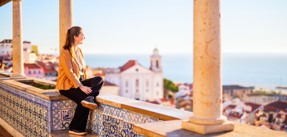 Woman in a yellow jacket sits on a tiled terrace, gazing at a scenic coastal town with a white church and red rooftops under a clear blue sky.