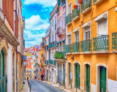 A colorful, narrow street in Lisbon with vibrant orange and yellow buildings, green balconies, cobblestone road, and a bright blue sky. Calm, picturesque vibe.