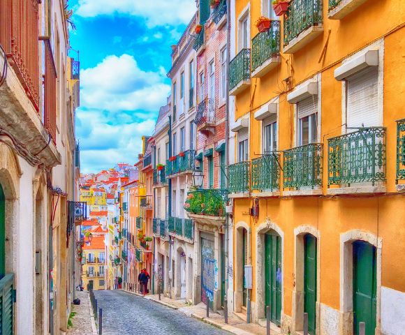 A colorful, narrow street in Lisbon with vibrant orange and yellow buildings, green balconies, cobblestone road, and a bright blue sky. Calm, picturesque vibe.