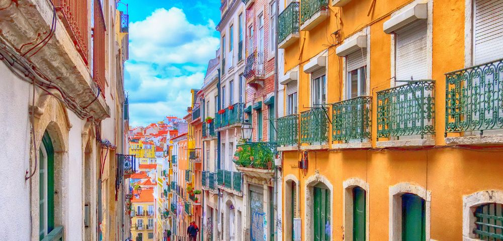 A colorful, narrow street in Lisbon with vibrant orange and yellow buildings, green balconies, cobblestone road, and a bright blue sky. Calm, picturesque vibe.