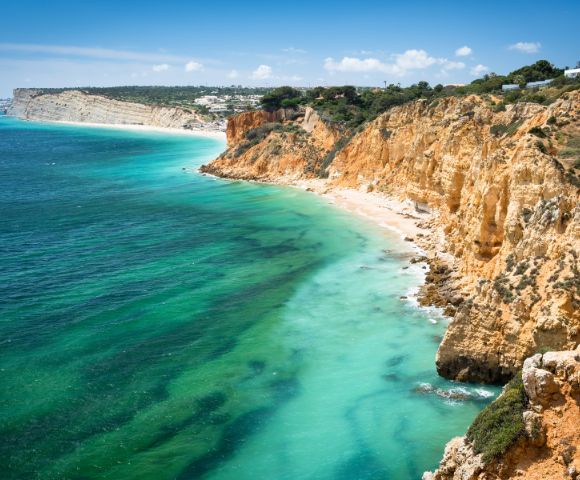 A stunning coastal landscape features rugged orange cliffs meeting turquoise sea, with clear skies above. The scene exudes tranquility and natural beauty.
