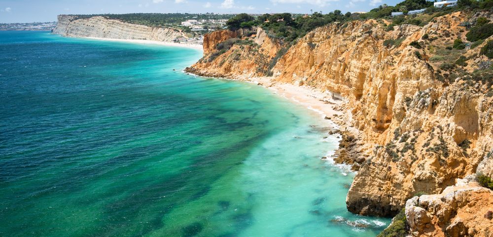 A stunning coastal landscape features rugged orange cliffs meeting turquoise sea, with clear skies above. The scene exudes tranquility and natural beauty.