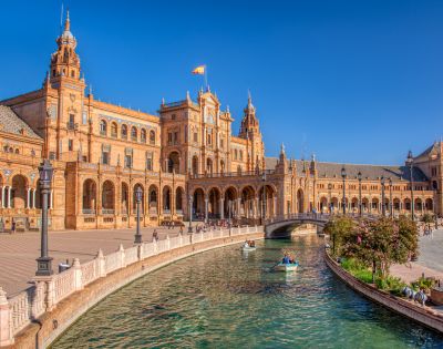Grand Spanish Renaissance Revival building with towers, arched walkways, and canal. People in boats, clear blue sky. Warm, elegant atmosphere.
