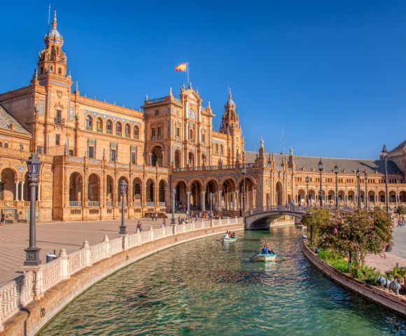 Grand Spanish Renaissance Revival building with towers, arched walkways, and canal. People in boats, clear blue sky. Warm, elegant atmosphere.