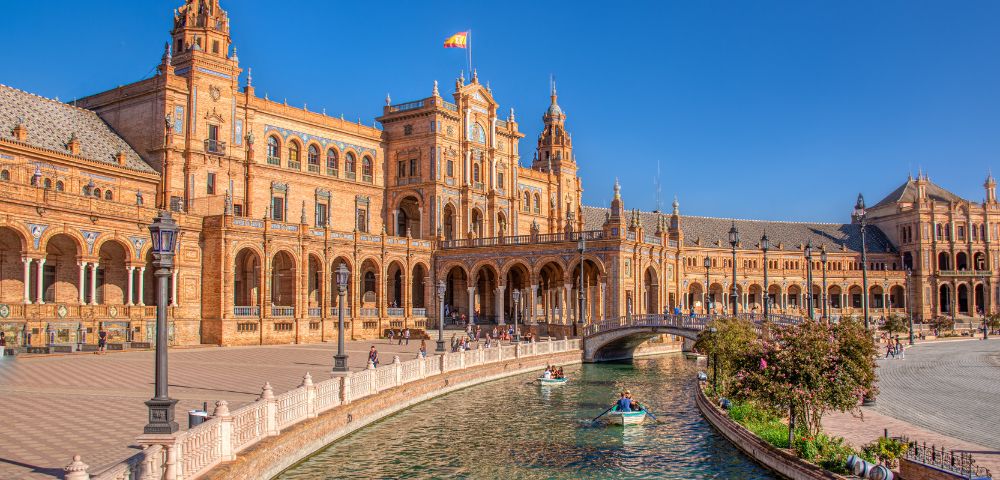 Grand Spanish Renaissance Revival building with towers, arched walkways, and canal. People in boats, clear blue sky. Warm, elegant atmosphere.