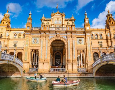 Grand architectural building with ornate columns and arches, surrounded by water. People in small boats, under a vibrant blue sky with fluffy clouds.