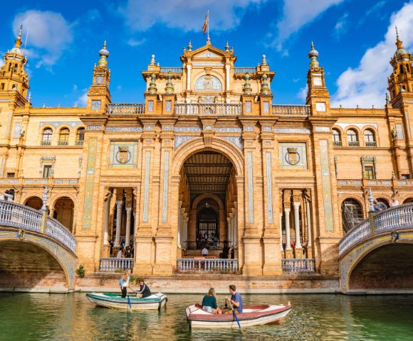 Grand architectural building with ornate columns and arches, surrounded by water. People in small boats, under a vibrant blue sky with fluffy clouds.