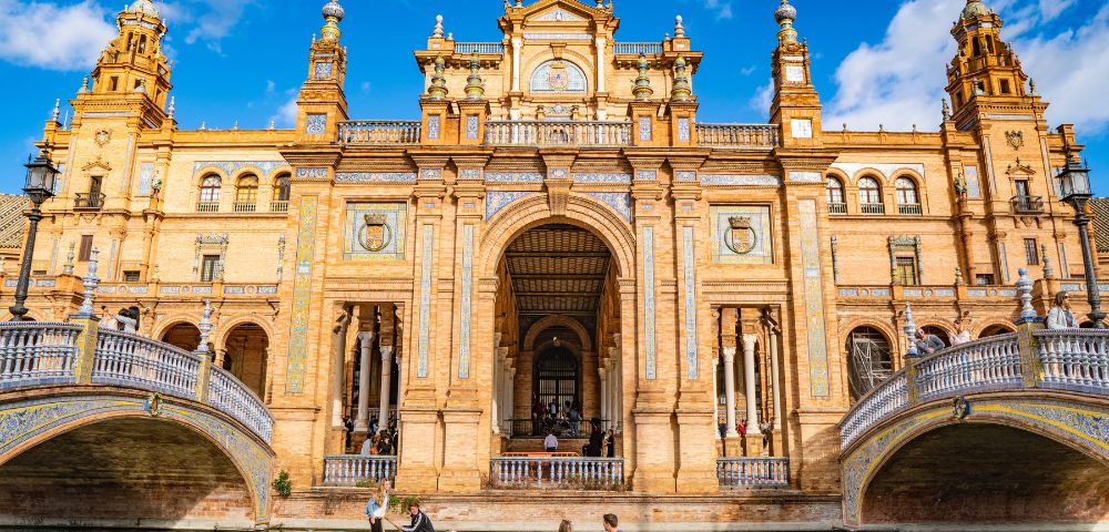 Grand architectural building with ornate columns and arches, surrounded by water. People in small boats, under a vibrant blue sky with fluffy clouds.