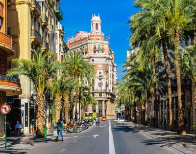Street scene in a sunny city lined with tall palm trees and classic European buildings. People and parked scooters create a lively urban atmosphere.