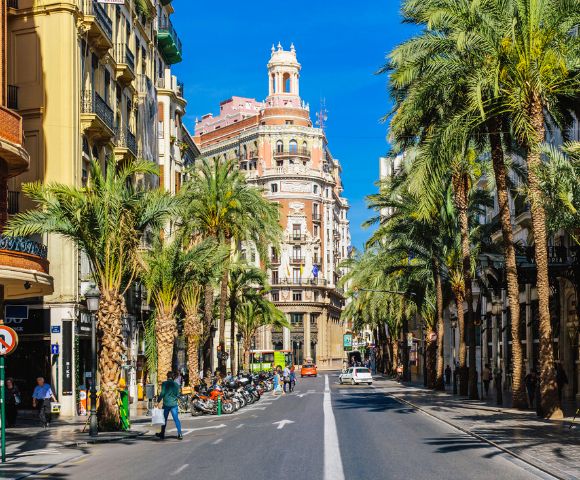 Street scene in a sunny city lined with tall palm trees and classic European buildings. People and parked scooters create a lively urban atmosphere.