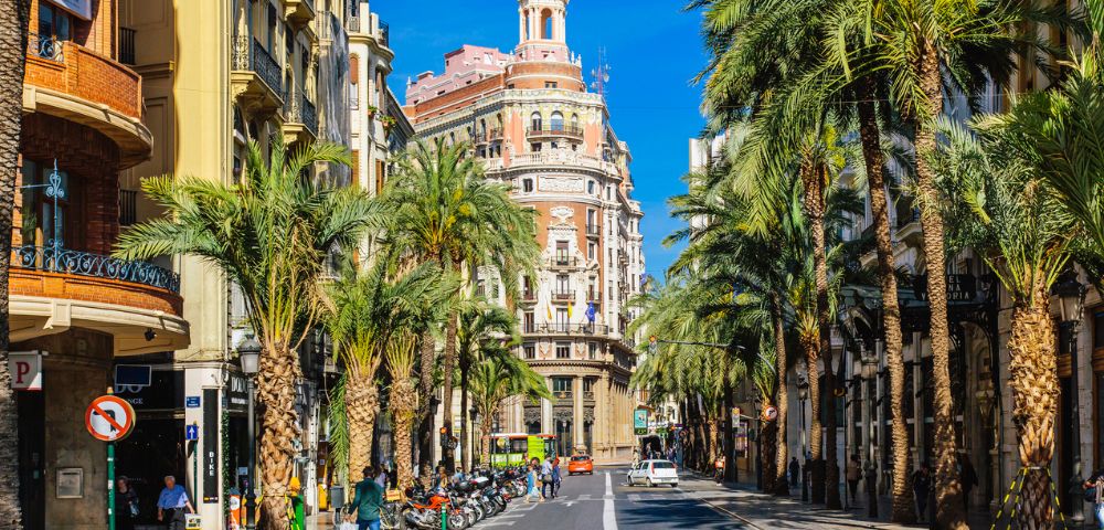 Street scene in a sunny city lined with tall palm trees and classic European buildings. People and parked scooters create a lively urban atmosphere.