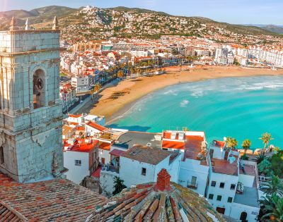 Seaside town with a sandy beach and turquoise water, bordered by colorful buildings and a bell tower. Hills stretch in the background under a clear sky.