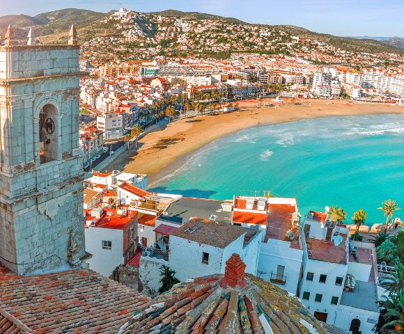 Seaside town with a sandy beach and turquoise water, bordered by colorful buildings and a bell tower. Hills stretch in the background under a clear sky.