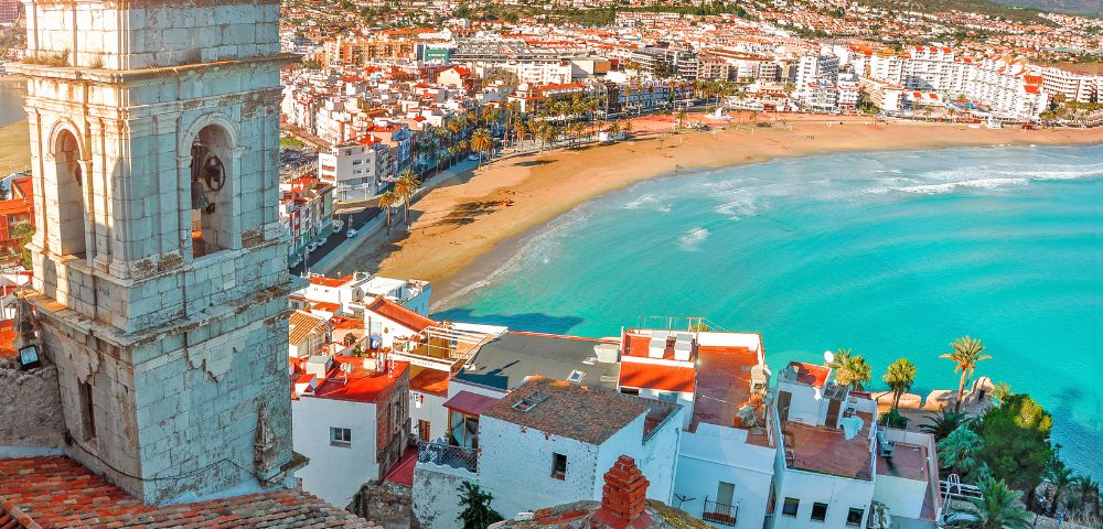 Seaside town with a sandy beach and turquoise water, bordered by colorful buildings and a bell tower. Hills stretch in the background under a clear sky.