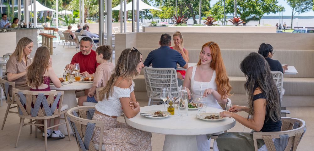 A group of people dine cheerfully at an outdoor restaurant with modern decor. Background shows lush trees and a sunny park, creating a relaxed atmosphere.