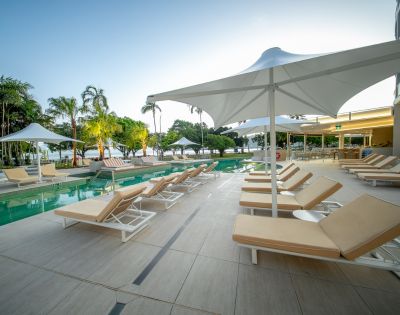 Poolside area with beige lounge chairs under white umbrellas on a tiled deck. Calm setting with trees and a clear sky in the background. Relaxing atmosphere.