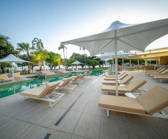 Poolside area with beige lounge chairs under white umbrellas on a tiled deck. Calm setting with trees and a clear sky in the background. Relaxing atmosphere.