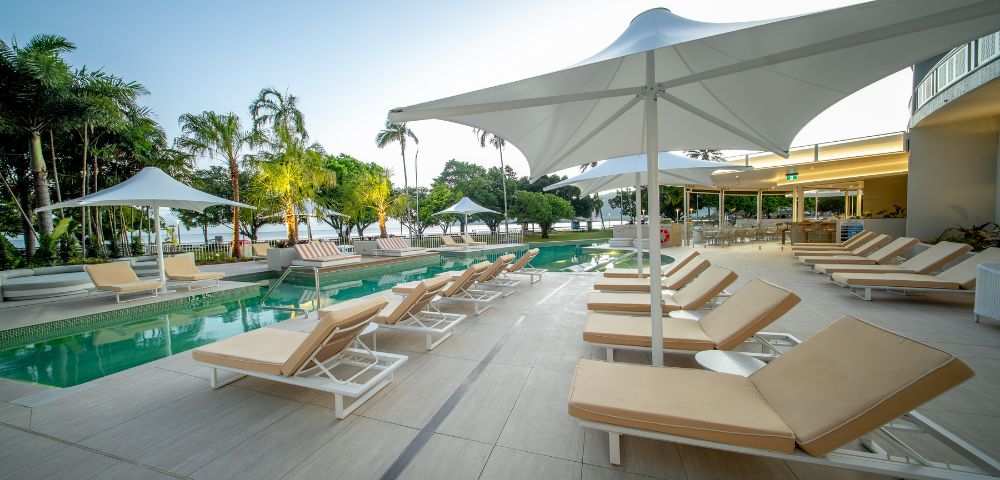 Poolside area with beige lounge chairs under white umbrellas on a tiled deck. Calm setting with trees and a clear sky in the background. Relaxing atmosphere.