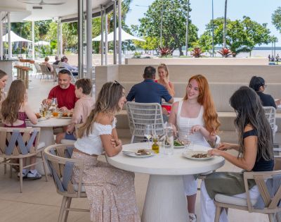 A group of people dine cheerfully at an outdoor restaurant with modern decor. Background shows lush trees and a sunny park, creating a relaxed atmosphere.