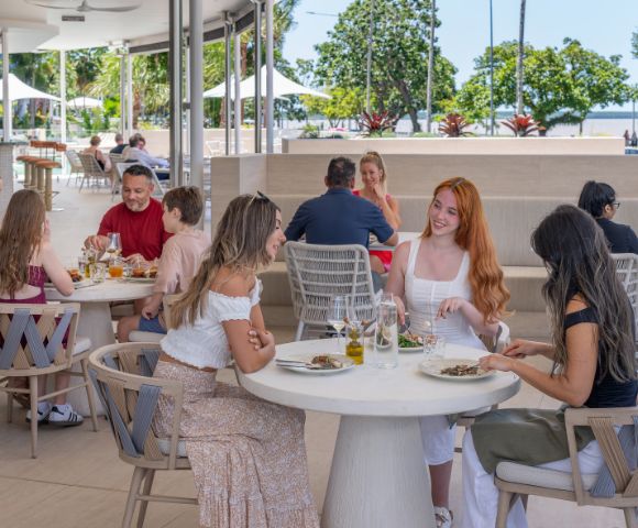 A group of people dine cheerfully at an outdoor restaurant with modern decor. Background shows lush trees and a sunny park, creating a relaxed atmosphere.