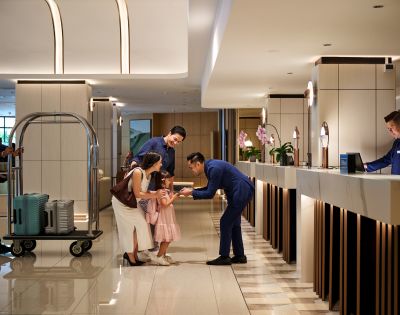 Hotel lobby scene with a smiling family checking in. A hotel staff member bends down to greet a young girl. A porter pushes a luggage cart. Warm, welcoming ambiance.