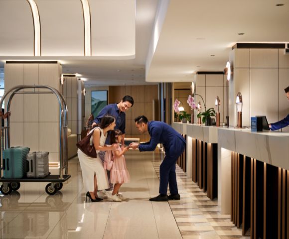 Hotel lobby scene with a smiling family checking in. A hotel staff member bends down to greet a young girl. A porter pushes a luggage cart. Warm, welcoming ambiance.