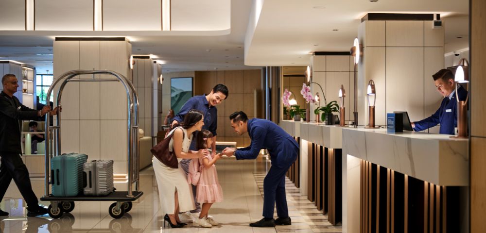 Hotel lobby scene with a smiling family checking in. A hotel staff member bends down to greet a young girl. A porter pushes a luggage cart. Warm, welcoming ambiance.