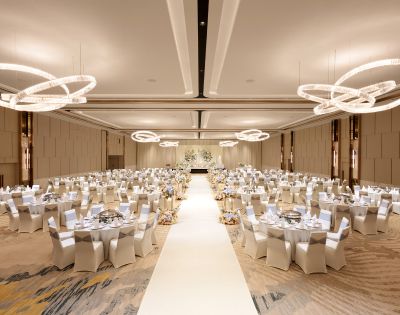 Elegant ballroom set for a wedding reception, featuring round tables with white chairs, a central aisle, and modern circular chandeliers, evoking a sophisticated ambiance.