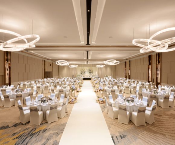 Elegant ballroom set for a wedding reception, featuring round tables with white chairs, a central aisle, and modern circular chandeliers, evoking a sophisticated ambiance.