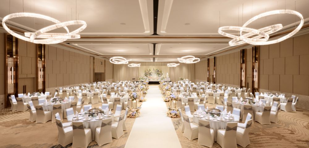 Elegant ballroom set for a wedding reception, featuring round tables with white chairs, a central aisle, and modern circular chandeliers, evoking a sophisticated ambiance.