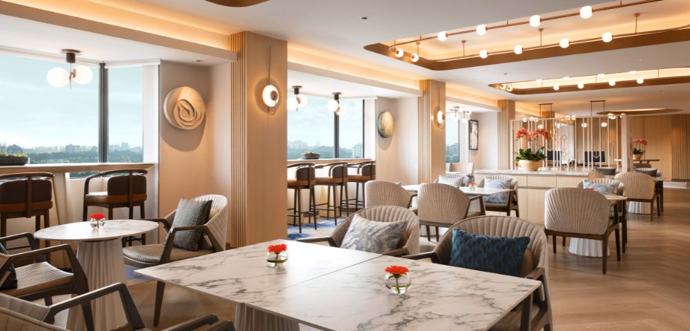 Stylish lounge with marble tables, modern chairs, and warm lighting. Large windows display a scenic view. Bright, inviting, and elegantly designed.