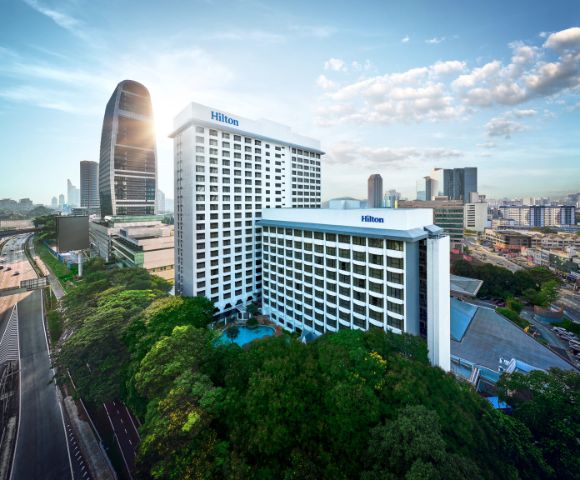 Aerial view of a Hilton hotel in a cityscape, with two white high-rise buildings surrounded by green trees, set against a skyline with modern skyscrapers under a partly cloudy sky.