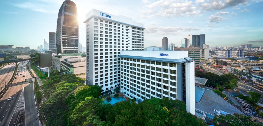 Aerial view of a Hilton hotel in a cityscape, with two white high-rise buildings surrounded by green trees, set against a skyline with modern skyscrapers under a partly cloudy sky.