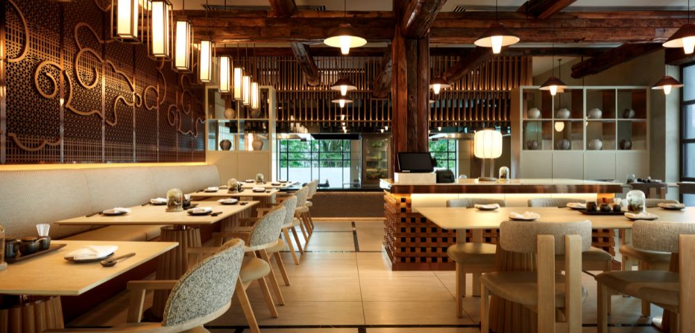 Elegant restaurant interior with wooden beams, warm lighting, and patterned walls. Tables are set for dining, creating a welcoming, cozy ambiance.