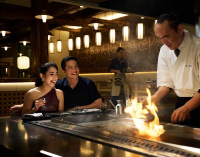 A couple cheerfully watches a chef cook on a hibachi grill in a dimly-lit restaurant, creating a lively and warm atmosphere with a fiery display.