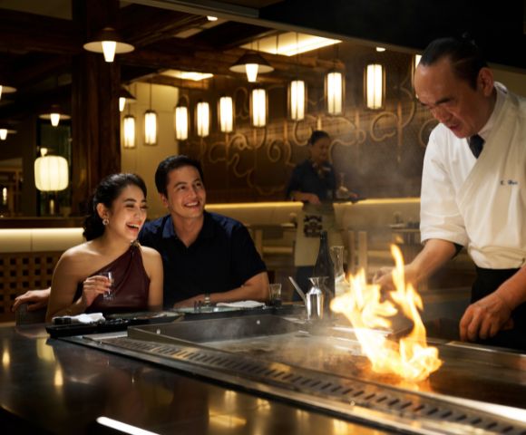 A couple cheerfully watches a chef cook on a hibachi grill in a dimly-lit restaurant, creating a lively and warm atmosphere with a fiery display.