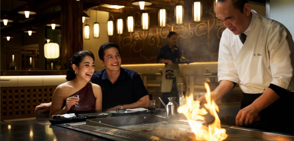 A couple cheerfully watches a chef cook on a hibachi grill in a dimly-lit restaurant, creating a lively and warm atmosphere with a fiery display.