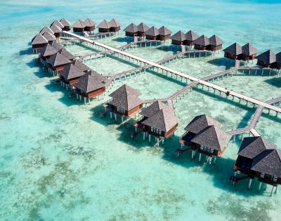 Aerial view of a serene tropical resort with multiple thatched-roof overwater bungalows connected by wooden walkways. Aqua-blue ocean conveys tranquility.