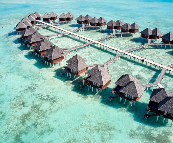Aerial view of a serene tropical resort with multiple thatched-roof overwater bungalows connected by wooden walkways. Aqua-blue ocean conveys tranquility.