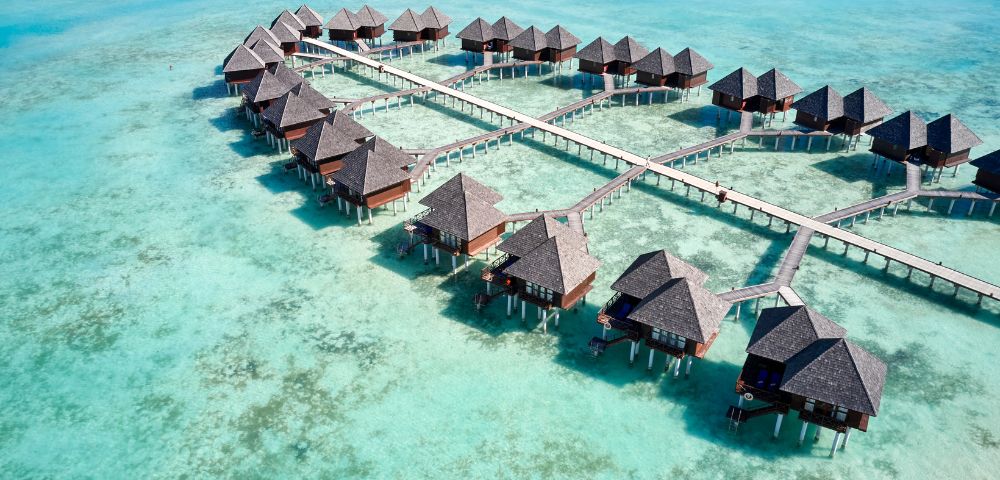 Aerial view of a serene tropical resort with multiple thatched-roof overwater bungalows connected by wooden walkways. Aqua-blue ocean conveys tranquility.