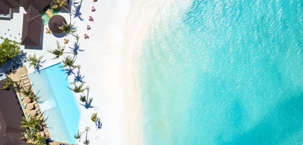 Aerial view of a tropical beach with turquoise water, white sand, palm trees, and beach umbrellas arranged in a line. Calm and serene atmosphere.