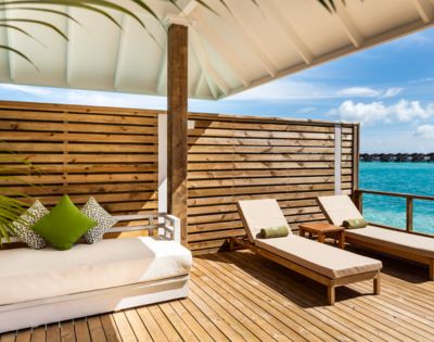 Sunlit deck overlooking turquoise ocean with two wooden lounge chairs and a cushioned sofa adorned with green pillows, framed by a wooden privacy screen.