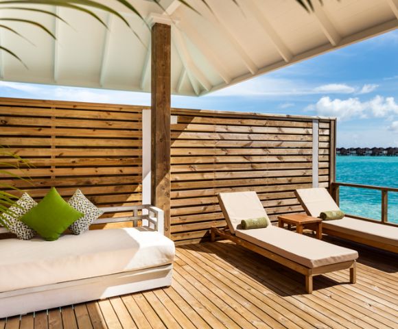 Sunlit deck overlooking turquoise ocean with two wooden lounge chairs and a cushioned sofa adorned with green pillows, framed by a wooden privacy screen.