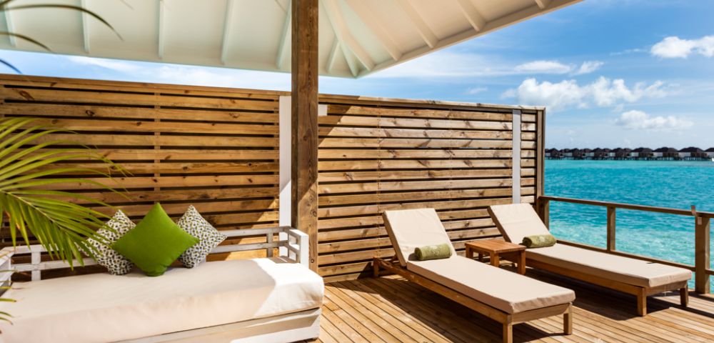 Sunlit deck overlooking turquoise ocean with two wooden lounge chairs and a cushioned sofa adorned with green pillows, framed by a wooden privacy screen.