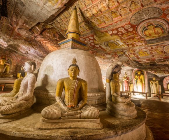 Ancient cave interior with serene stone Buddha statues in meditation poses surrounding a central stupa, adorned with vibrant, detailed ceiling murals.