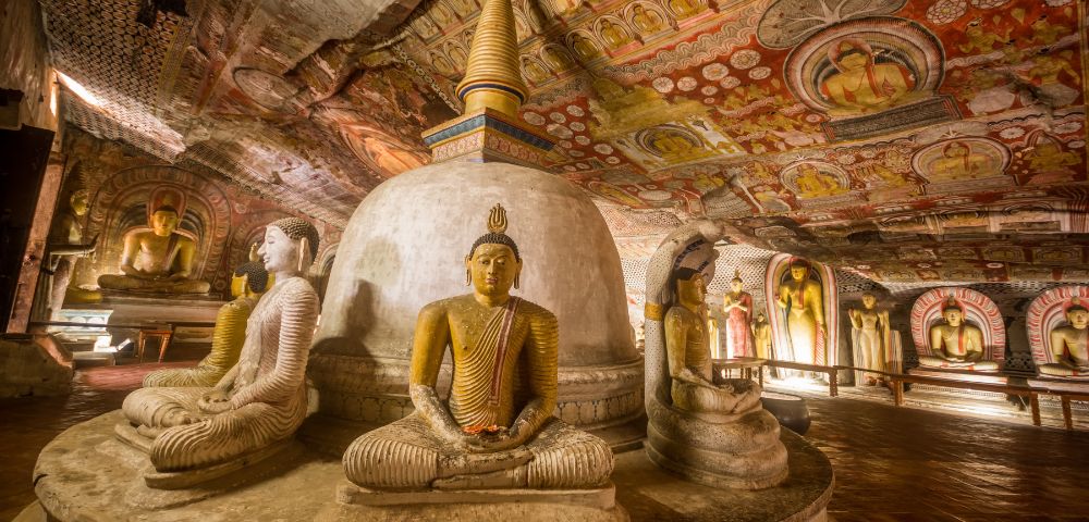 Ancient cave interior with serene stone Buddha statues in meditation poses surrounding a central stupa, adorned with vibrant, detailed ceiling murals.