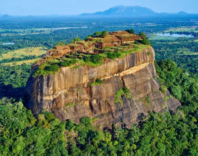 Aerial view of Sigiriya, Sri Lanka, showcasing the ancient rock fortress atop a massive rock formation, surrounded by lush green forest and distant mountains.
